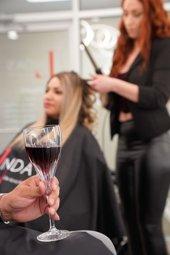 A male client holds a glass of red wine while seated in a salon, a female client gets her hair styled in the background